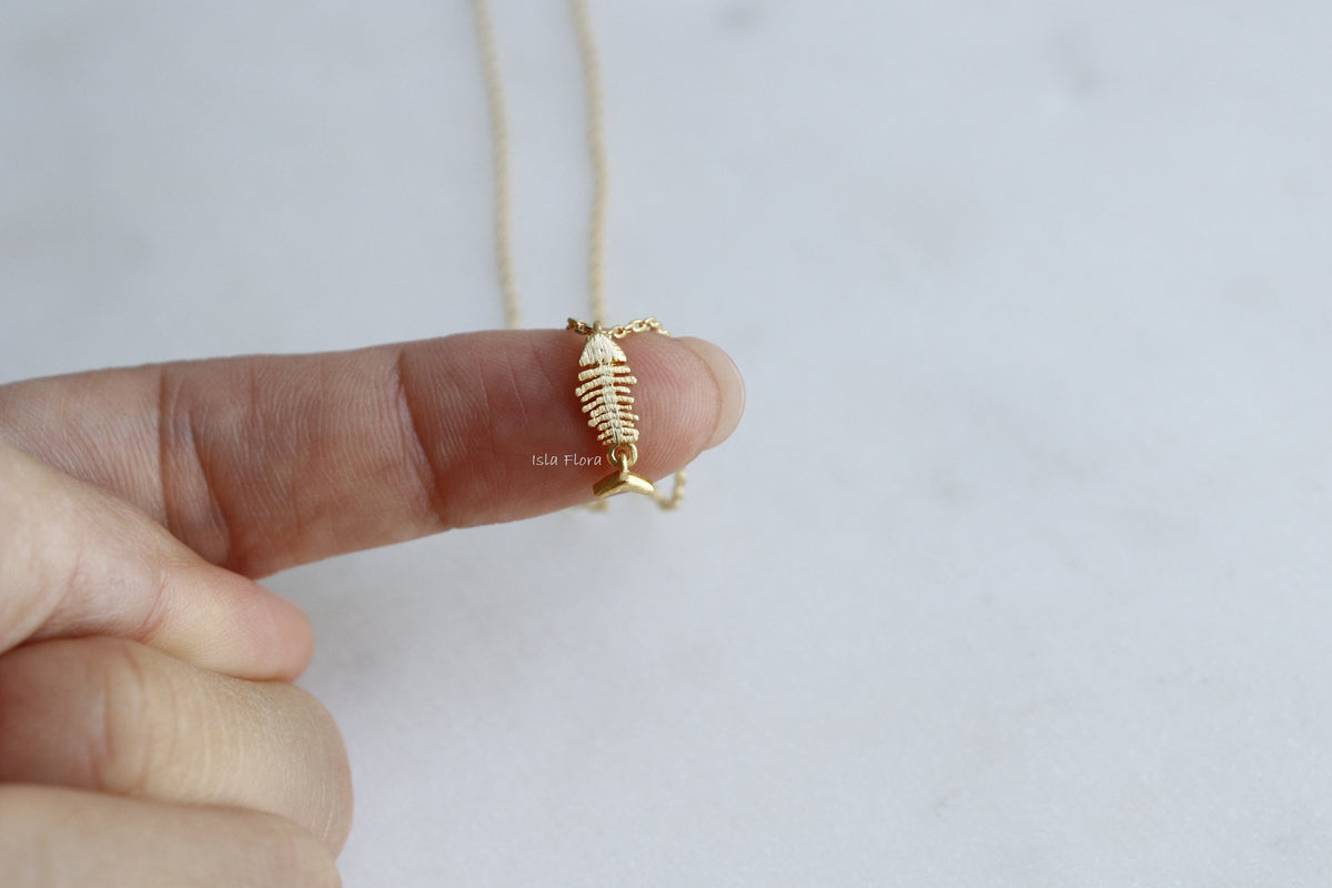 A gold fish skeleton pendant necklace held on a person's finger against a light gray background.