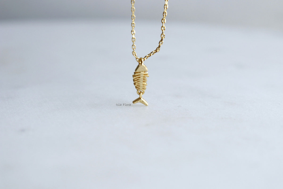 isla flora A delicate gold necklace featuring a small fish skeleton pendant, suspended against a soft, light gray background.