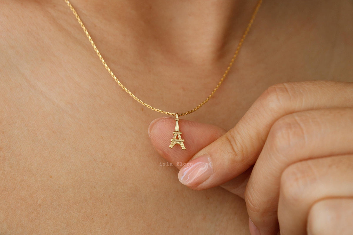 A close-up of a delicate gold chain necklace with a small Eiffel Tower pendant, worn on a person's neck with a hand gently touching the pendant.