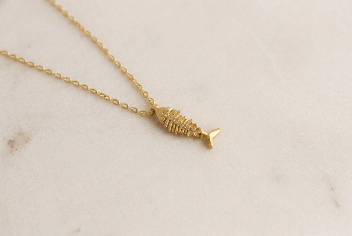 A delicate gold chain necklace featuring a small fish skeleton pendant with a small gold fin, displayed on a light gray textured surface.