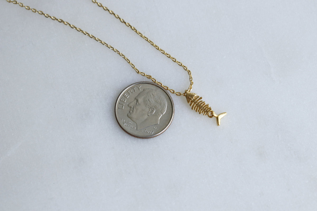 A gold chain necklace featuring a small gold fish skeleton pendant next to a US dime for scale, displayed on a light gray background.