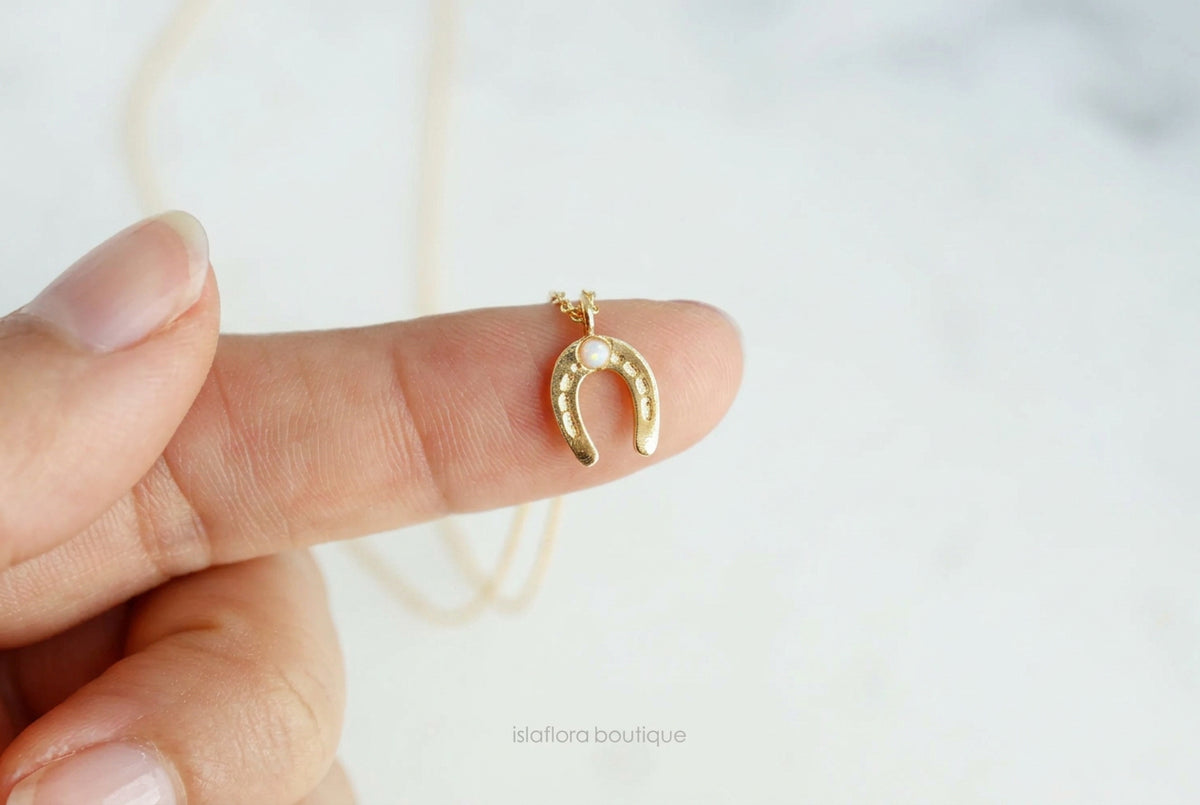 Gold horseshoe-shaped pendant on a chain held between fingers against a light background.