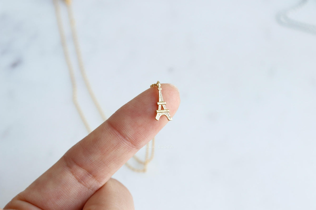 A gold Eiffel Tower pendant necklace held on a person's finger against a light marble background.