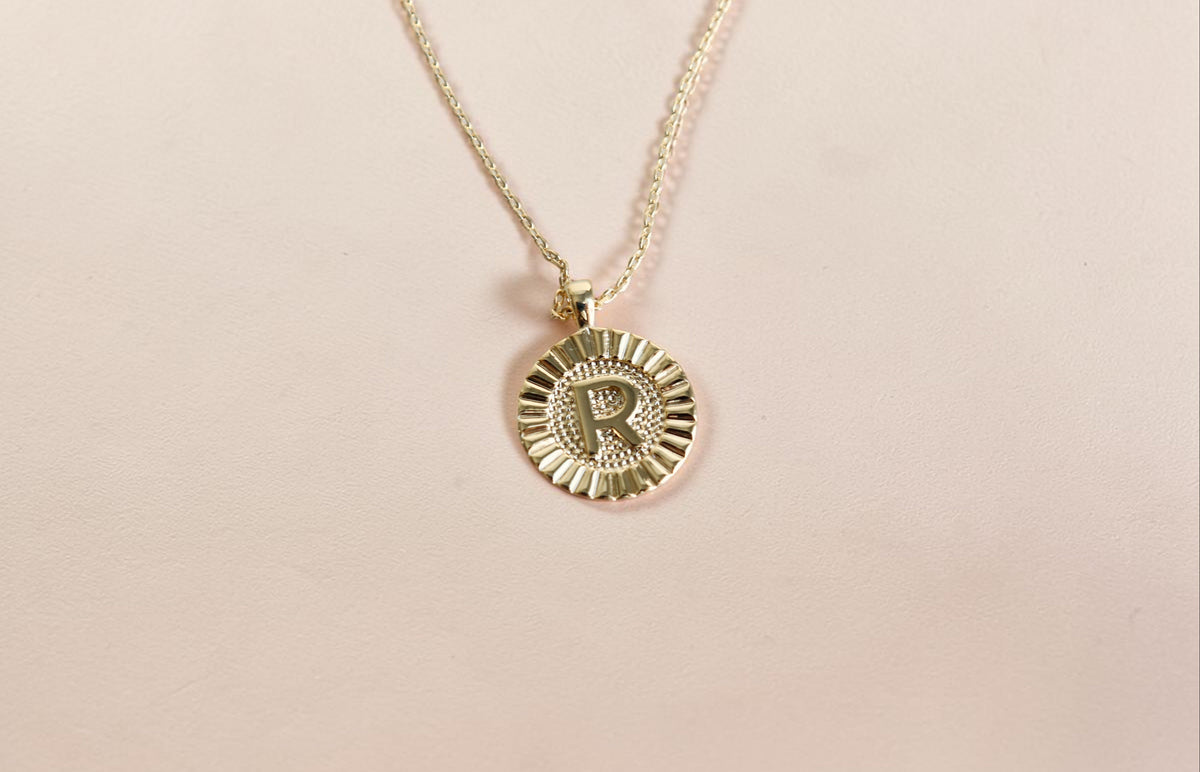 A gold initial necklace featuring a circular sunburst pendant with the letter 'R' engraved in the center, suspended from a delicate gold chain against a solid pale pink background.
